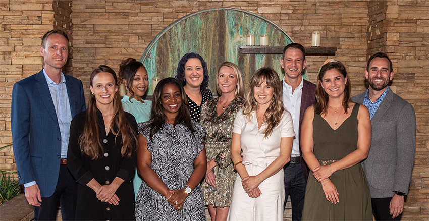 A group of eleven adults, dressed in business casual attire, stand together smiling in front of a stone wall with a circular green metal accent and candles, representing the Weaver Philanthropic Initiative.