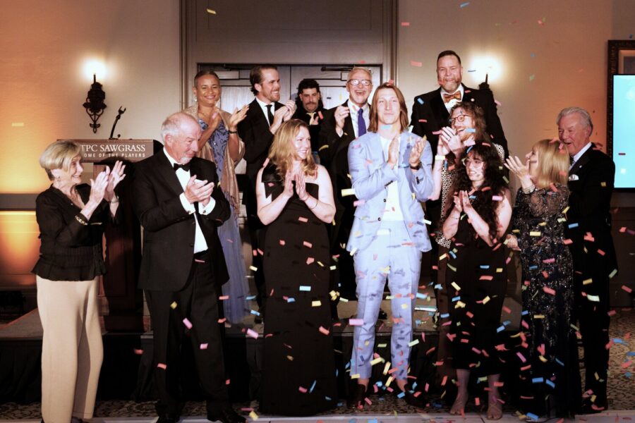 A group of people dressed in formal attire stand on a stage clapping and smiling as colorful confetti falls around them, celebrating a fundraising record at the Gala for Here Tomorrow.