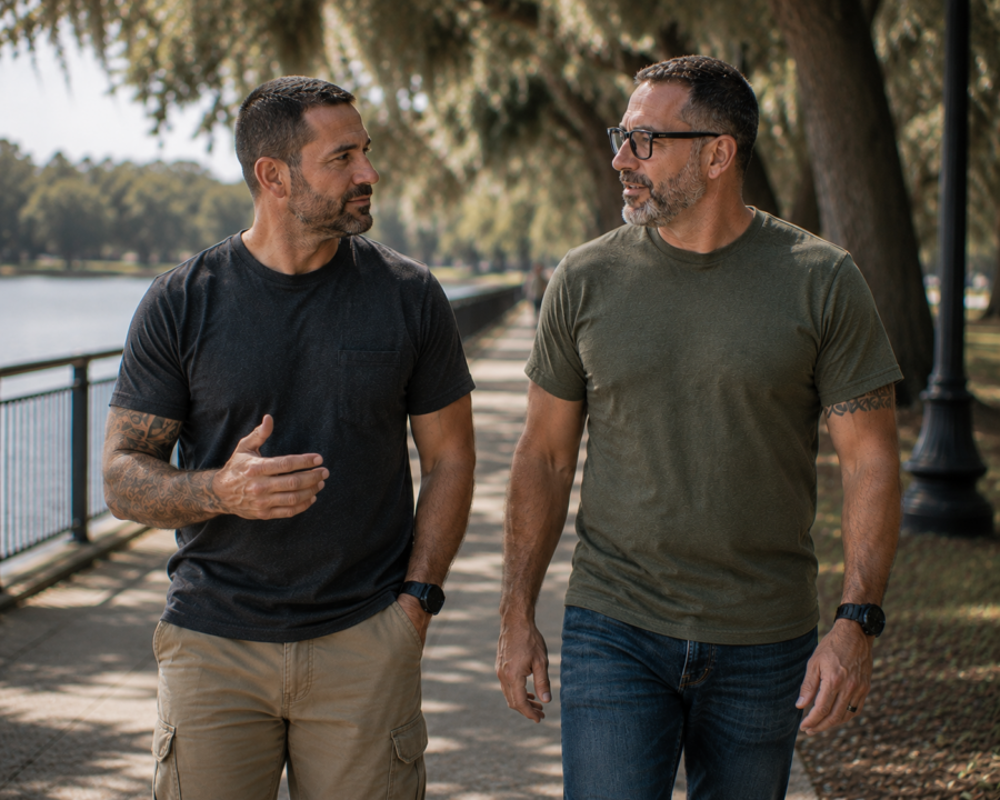 Two men with short hair and beards walk and talk along a tree-lined path beside a body of water on a sunny day, their conversation touching on suicide prevention resources and the importance of supporting one another.
