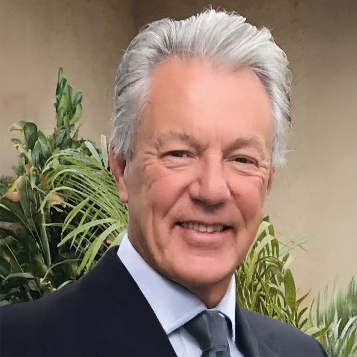 Smiling older man with gray hair wearing a suit and tie, standing outdoors in front of green plants—Chris Scangas.