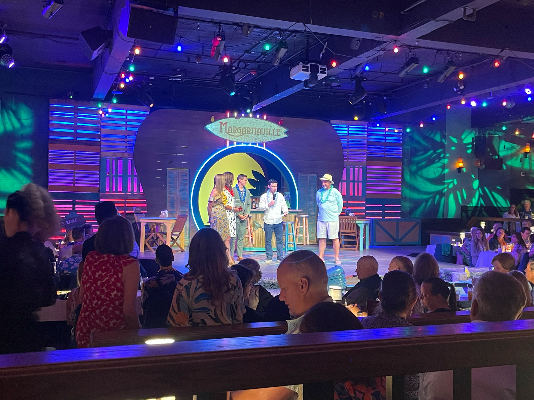 A group of people stands on a stage under colorful lights, with an audience seated at tables watching the event in a lively indoor venue.