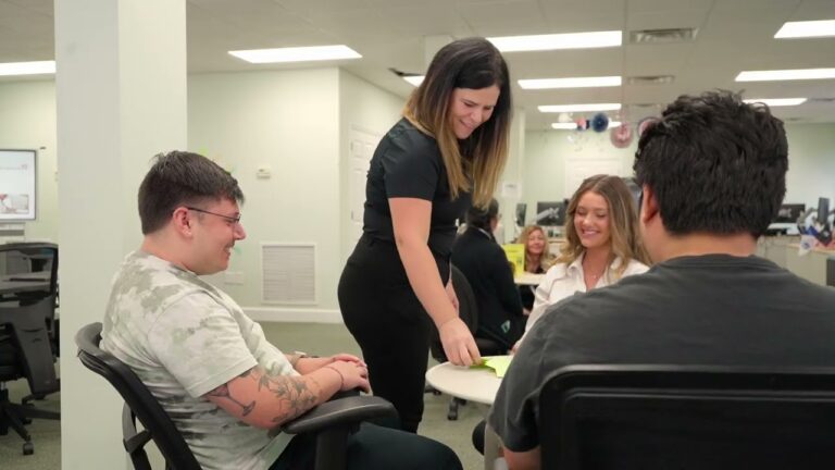 Four people sit around a table in an office as one woman stands, handing papers to a seated man—perhaps discussing Peer Support Certification. Other desks and people are visible in the background.