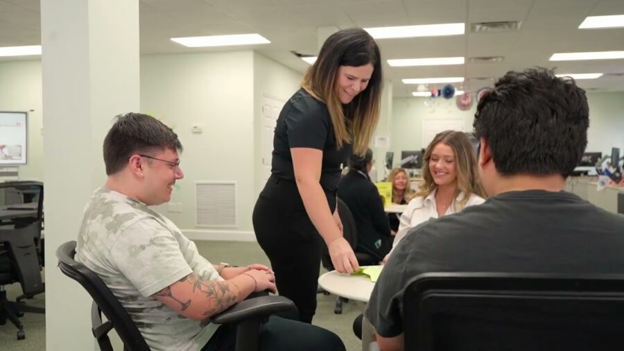 Four people sit around a table in an office as one woman stands, handing papers to a seated man—perhaps discussing Peer Support Certification. Other desks and people are visible in the background.