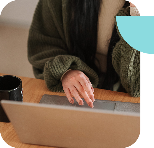 Person wearing a green sweater using a laptop at a wooden table with a black mug nearby. The image is partly overlaid with blue and white geometric shapes.
