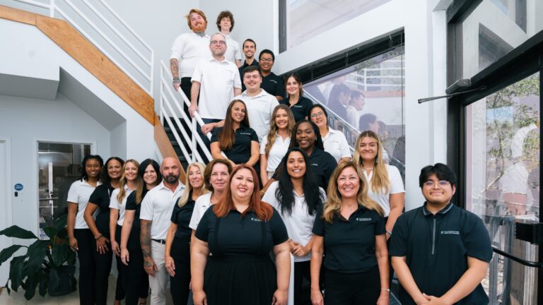 A group of twenty people, dressed in black and white business casual attire, pose together inside a modern office near a staircase and large windows for a folio weekly feature.