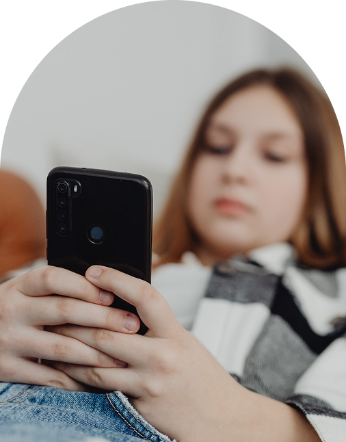 A young person holding and looking at a smartphone while lying down, with the phone in focus and their face slightly blurred.