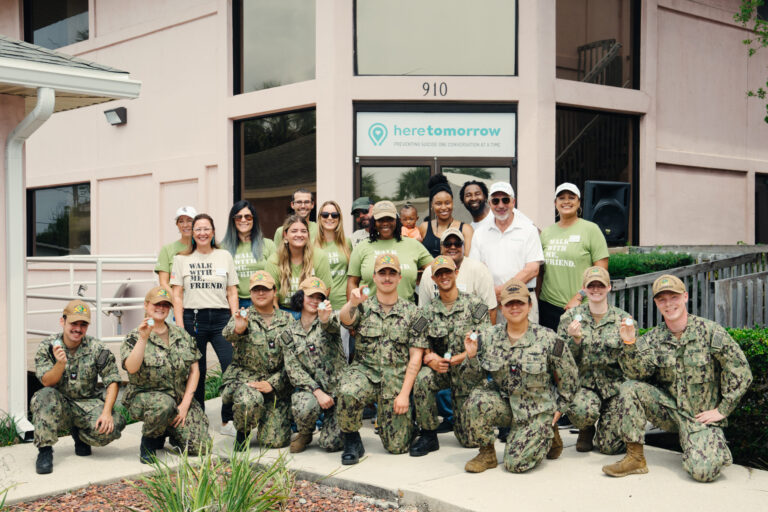 A group of people, some in military uniforms and others in casual clothes, pose together outside a building with a "heretomorrow" sign during an honor gathering.