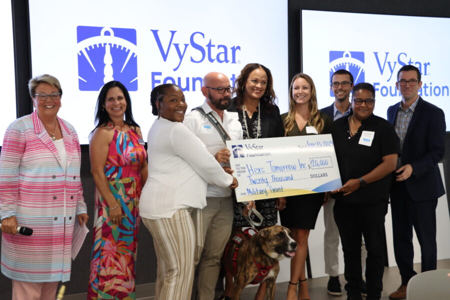 A group of people stands together indoors, holding a $20,000 grant check from VyStar Foundation to Heroes Tomorrow Inc. Two display screens show the VyStar Foundation logo, highlighting support for the Military Peer Support Program.