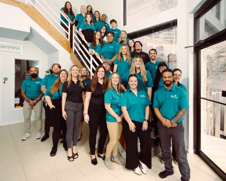 Meet our team: a group of people wearing teal shirts and black or khaki pants pose for a photo on and around a staircase in a bright, modern office setting.